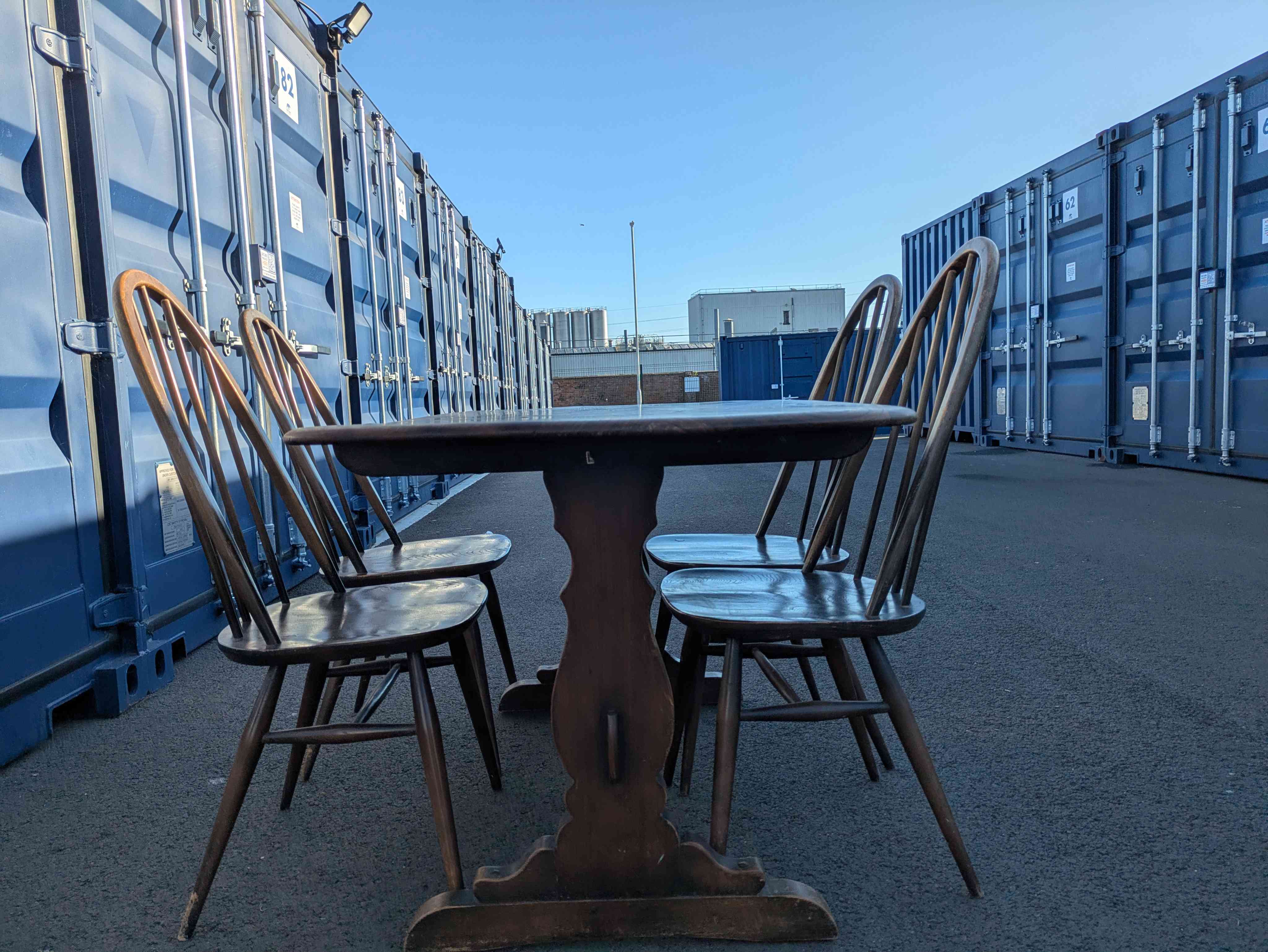 A small dark oak refectory table with two spindle-back chairs, photographed in the sun between two storage containers